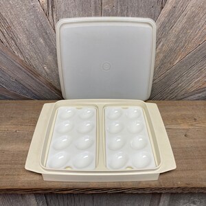 May include: A vintage off-white plastic egg container with a matching lid. The container has two sections, each holding a dozen egg-shaped indentations. The lid is square with a slightly rounded edge. The container is open, revealing the egg holders.