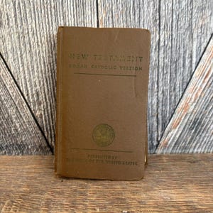 May include: A brown, vintage New Testament book with the text "New Testament Roman Catholic Version" in green lettering. The book is presented by the Army of the United States.