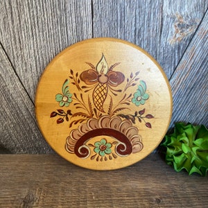 May include: A round wooden plaque with a hand-painted floral design. The design features brown, yellow, and teal flowers with brown and green leaves. The plaque is perfect for adding a touch of rustic charm to any home.