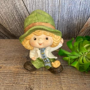 May include: A ceramic figurine of a child wearing a green hat and a blue and white scarf. The child is holding a small yellow bird figurine. The figurine is in a sitting position and has a cheerful expression.