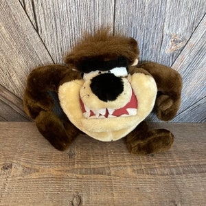 May include: A plush toy of the Tasmanian Devil, featuring brown fur, a black nose, and a wide, toothy grin. The toy has large arms and feet, and a tuft of brown hair on top of its head. The character is set against a wooden background.