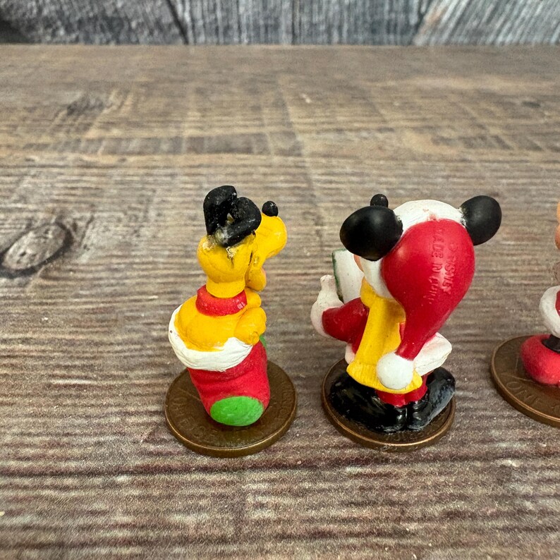 May include: A collection of small, colorful Disney character figurines. One figure is a yellow dog with black ears and a red collar. Another is Mickey Mouse in a Santa hat and red coat. The figurines are on bronze-colored bases.