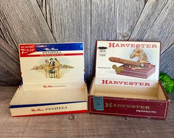 May include: Two vintage cigar boxes. The box on the left is blue, white, and red with the text "Wm. Penn PANATELA THE GREAT AMERICAN CIGAR" and "Wm. Penn PANATELA". The box on the right is red and white with the text "HARVESTER PERFECTO" and a horse head.