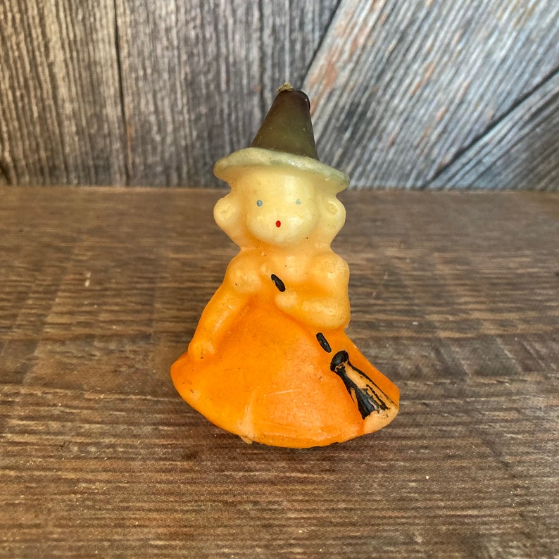 May include: A vintage Halloween candle shaped like a witch with a pointed hat, orange dress, and a black broom. The candle is made of wax and has a painted face with blue eyes and a red nose.