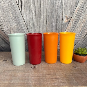 May include: Four colorful plastic tumblers in shades of mint green, red, orange, and yellow are arranged on a wooden surface. The tumblers are cylindrical with a slightly flared top. A small potted succulent is to the right.