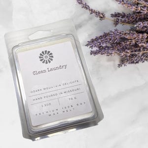 Puede incluir: Un paquete de plástico transparente que contiene una cera perfumada blanca con el texto "Clean Laundry". La etiqueta también dice "Ozark Mountain Delights" y "Hand Poured in Missouri". La cera perfumada pesa 70 g. Una ramita de lavanda púrpura está en el fondo.