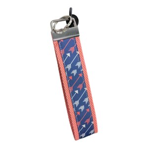 Può includere: Un portachiavi rosa corallo con un motivo a frecce blu e bianche. Il portachiavi ha un anello in metallo argentato.