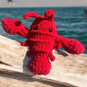 May include: A red crocheted lobster plush toy with black eyes. The toy is sitting on a wooden surface with a blue ocean in the background.