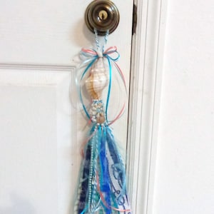 May include: A decorative hanging ornament featuring a spiral seashell at the top, adorned with small white beads and shells. Below, a collection of blue, white, and coral ribbons and fabric strips hang down, tied with a matching blue and white cord.