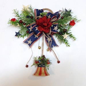 May include: A patriotic wreath with a red, white, and blue bow, red flowers, and a bell with a red, white, and blue design. The wreath is decorated with green leaves, white stars, and red berries.