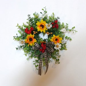 May include: A sunflower wreath with yellow, red, and white flowers, green leaves, and a black and white checkered ribbon.
