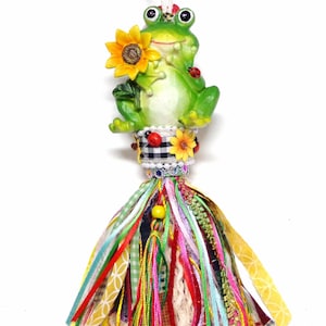 May include: A whimsical green frog figurine holding a yellow sunflower, adorned with ladybugs and a colorful tassel. The frog is perched atop a decorative element with a black and white checkered pattern, perfect for a garden or nature-themed decor.