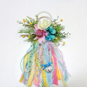 May include: A decorative floral hanging ornament with a white rope loop. It features a cluster of colorful artificial flowers in pink, teal, and white, accented with greenery. Below, colorful ribbons and a small bell with a blue bow dangle.