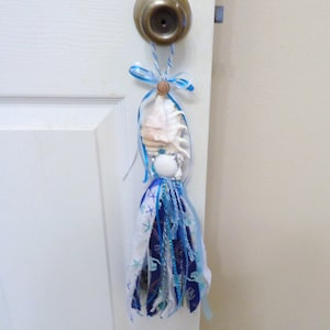 May include: A blue and white fabric tassel with a seashell and beads hanging from a door knob. The tassel is decorated with a blue and white fabric with a starfish pattern.