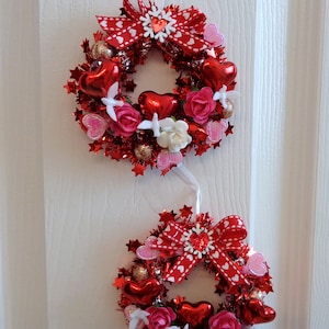 May include: Two small, decorative Valentine's Day wreaths. Each wreath is red with heart and star accents, red bows, and small pink and white flowers. They hang from white ribbons.
