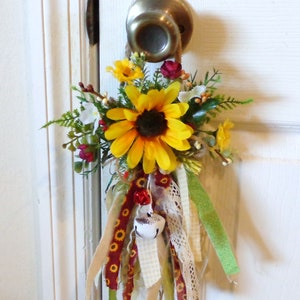 May include: A decorative door hanger featuring a large yellow sunflower with a dark center, surrounded by smaller yellow and red flowers, and greenery. The hanger includes ribbons in various colors and patterns, and a small bell.
