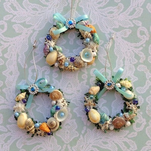 May include: Three handmade Christmas ornaments shaped like wreaths, decorated with seashells, blue and white beads, and blue ribbon bows.