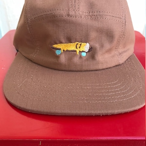 Peut inclure: Une casquette marron à 5 panneaux avec un motif brodé jaune et bleu représentant un bonhomme qui fait du skateboard.