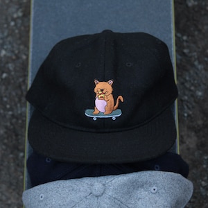 May include: Black baseball cap with a cartoon cat skateboarding and eating a sandwich embroidered on the front.