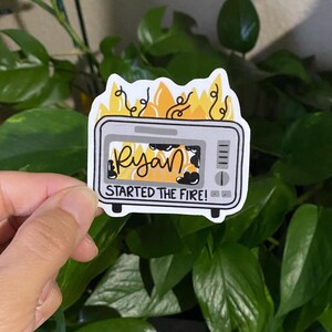 May include: A white sticker featuring a cartoon toaster engulfed in flames. The toaster is gray with black accents and the text "Ryan STARTED THE FIRE!" The flames are yellow and orange. The sticker is held in front of a background of green leaves.