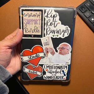 May include: A black Kindle e-reader with various stickers, including a heart with the word "Slade" on it, a heart with the word "Kaden" on it, a sticker with the text "Emotional Support Kindle", a sticker with the text "Rip Rot Ravinger", a sticker with the text "Emotionally attached to the Wingleader", and a sticker with the text "Fourth Wing".