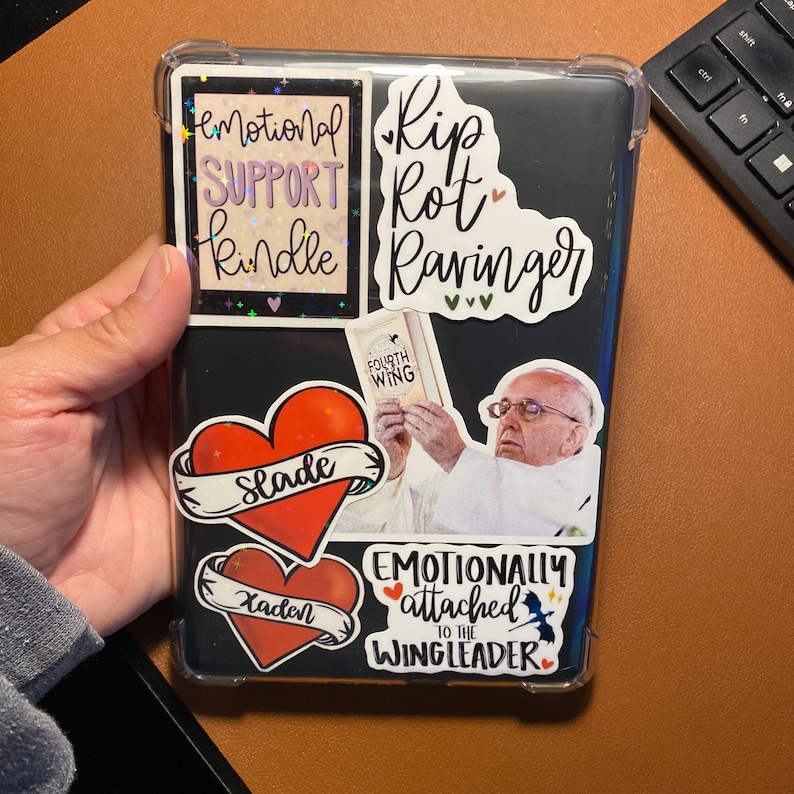 May include: A black Kindle e-reader with various stickers, including a heart with the word "Slade" on it, a heart with the word "Kaden" on it, a sticker with the text "Emotional Support Kindle", a sticker with the text "Rip Rot Ravinger", a sticker with the text "Emotionally attached to the Wingleader", and a sticker with the text "Fourth Wing".