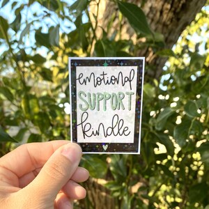 May include: A holographic sticker with a black background and white stars. The sticker says "emotional support kindle" in green and black lettering.