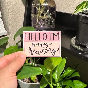 May include: A rectangular sticker with the text "HELLO I'M busy reading" in a handwritten style. The sticker has a pink and white background with glitter accents. The sticker is held in front of green plants.