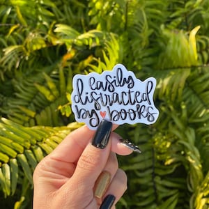 May include: A white sticker with black script that reads "easily distracted by books" with a small red heart. The sticker is held in front of a background of green foliage.