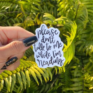 May include: A white sticker with black text that reads "please don't talk to me while I'm reading." The sticker is held in front of a background of green foliage.