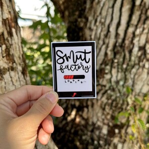May include: A rectangular sticker with a black border features the words "Smut Factory" in cursive above a loading bar graphic. The loading bar is red and black. A small red "S" is at the bottom.
