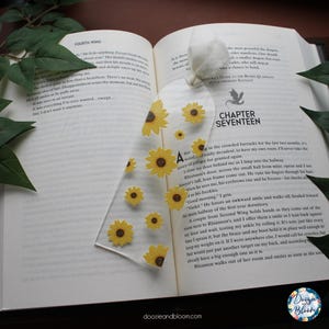 May include: A clear acrylic bookmark featuring yellow sunflower designs. The bookmark is rectangular with rounded corners and is placed on an open book. The bookmark is a decorative accessory for readers.