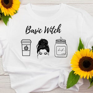 May include: White t-shirt with the text "Basic Witch" printed in black. The design includes a pumpkin spice latte, a woman with a messy bun, and a candle.