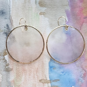 May include: A pair of circular drop earrings featuring thin gold-toned metal rims surrounding translucent, iridescent discs with a subtle pearlescent sheen. Each earring is suspended from a simple gold-toned wire hook for attachment.