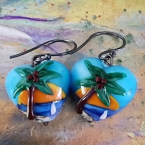 May include: A pair of heart-shaped glass earrings featuring a tropical scene with a green palm tree, a bright orange sun, and a blue ocean background. The earrings are suspended from dark metal hooks, showcasing a detailed, colorful design.