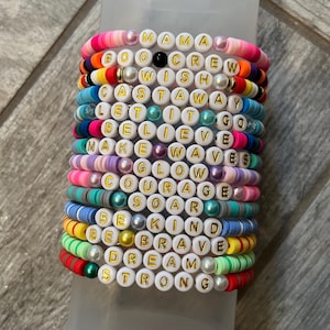 May include: Stack of colorful beaded bracelets with white letter beads spelling out words like "MAMA", "CREW", and "DREAM". The bracelets are in various colors including pink, blue, green, and yellow. The bracelets are stacked on top of each other.