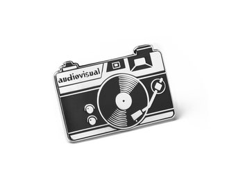 Pode incluir: Pin de esmalte preto e branco de uma câmera com um toca-discos dentro. A câmera tem a palavra "audiovisual" impressa nela.