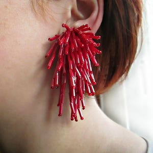 Könnte beinhalten: Ein leuchtend roter Perlenohrring bestehend aus einem dichten Büschel langer, zylindrischer Glasperlen, die in einer fransenartigen Anordnung nach unten hängen und ein strukturiertes, mehrschichtiges Erscheinungsbild erzeugen, das das Licht einfängt.