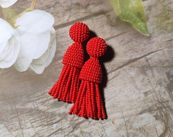 May include: A pair of vibrant red beaded tassel earrings. Each earring has a spherical top and a long, fringed tassel. The earrings are presented on a wooden surface with floral details in the background.