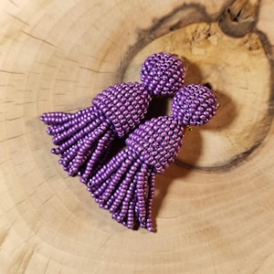 May include: A pair of beaded tassel earrings in a vibrant purple hue. The earrings feature a round, beaded top and a long, fringed tassel design. The earrings are displayed on a wooden surface.