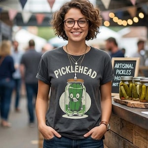 May include: A charcoal grey short-sleeve t-shirt featuring a graphic of a cartoon pickle inside a glass jar with the text PICKLEHEAD printed above it. The shirt is paired with dark blue denim shorts with rolled cuffs and a brown leather watch.