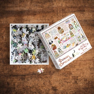 May include: A 1000 piece jigsaw puzzle featuring a colorful illustration of a cat reading a book, surrounded by books and other literary objects. The puzzle box is white with a red and black border and the text "Susan Branch For the Love of Books 27"x20" 1000 Piece Puzzle".