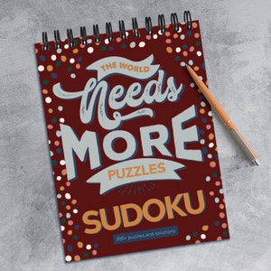 May include: A red spiral bound notebook with a colourful polka dot pattern. The cover features the text "The World Needs More Puzzles Sudoku" with the word "Sudoku" in orange. The text "200+ puzzles and solutions" is printed at the bottom of the cover.