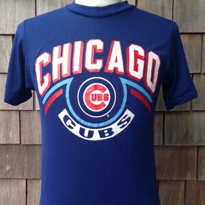 May include: A blue t-shirt with a white, red, and blue Chicago Cubs logo. The logo features the word "Chicago" in a white and red arc above a circle with the word "Cubs" in a white arc below. The circle contains a red "C" with a white "UBS" inside.