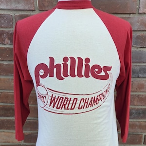 May include: A cream-colored baseball-style shirt with red three-quarter length sleeves and a red collar. The front features a red graphic with the text 'phillies' above a baseball and the words '1980 WORLD CHAMPIONS' in a curved design.