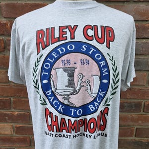 May include: A gray t-shirt featuring a graphic commemorating the Toledo Storm hockey team's Riley Cup wins in the 1992-93 and 1993-94 seasons. The design includes the words "Riley Cup," "Toledo Storm," and "Champions."