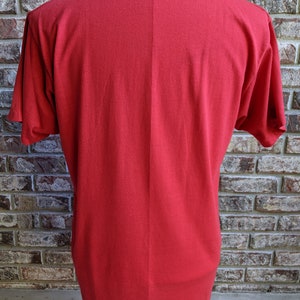 May include: A solid red t-shirt with short sleeves. The back of the shirt is displayed, showing the neckline and the fabric's texture. The shirt is on a mannequin against a brick wall background.
