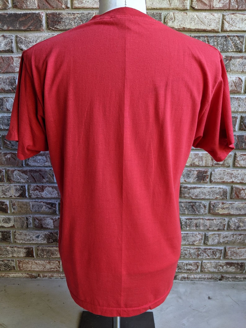 May include: A solid red t-shirt with short sleeves. The back of the shirt is displayed, showing the neckline and the fabric's texture. The shirt is on a mannequin against a brick wall background.