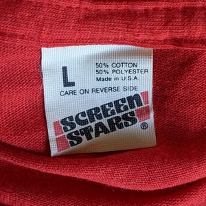 May include: Close-up of a red t-shirt with a white tag. The tag reads "L", "50% COTTON 50% POLYESTER Made in U.S.A." and "CARE ON REVERSE SIDE". The tag also features the "SCREEN STARS" logo.