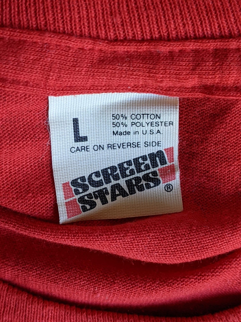 May include: Close-up of a red t-shirt with a white tag. The tag reads "L", "50% COTTON 50% POLYESTER Made in U.S.A." and "CARE ON REVERSE SIDE". The tag also features the "SCREEN STARS" logo.